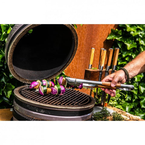 Kamado kepsninė atidaryta su dangčiu; ranka laiko medinėmis rankenomis nerūdijančias žnyples, ant grotelių kepami spalvoti daržovių iešmeliai